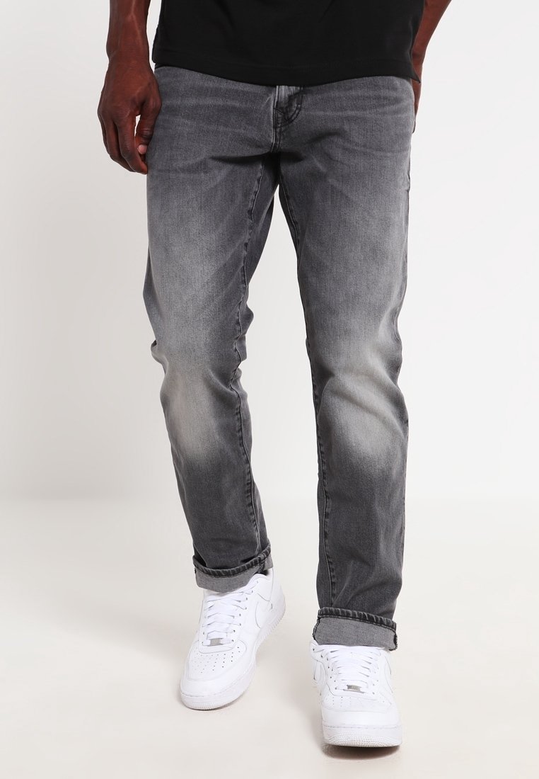 Homme portant un jean gris délavé avec les ourlets retroussés et des baskets blanches, debout devant un fond clair uni.