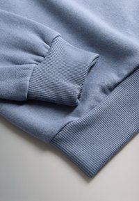 Gros plan sur la manche d'un sweat-shirt bleu clair avec un poignet côtelé et une ceinture côtelée assortie sur une surface claire.