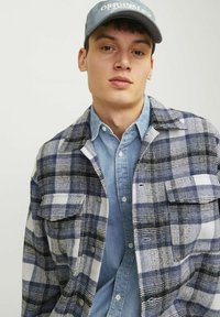 Rutete flannel skjorte i marineblå og hvid med lommer; båret over en lyseblå denim skjorte. Kappen er grå med hvid tekst. Tekstureret stof.