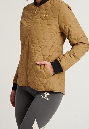 Persoon draagt een beige doorgestikte jas met rits en zwarte manchetten, en grijze leggings met witte hummel-logo's, hand in de jaszak.