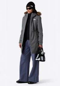 Chaqueta larga gris con capucha forrada de piel, combinada con pantalones anchos de pierna azul marino. Gorro negro y gafas de sol; sosteniendo una bolsa en blanco y negro.