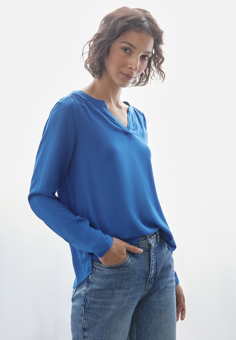 Blusa blu a maniche lunghe con scollatura a V e spacchi laterali. Indossata con jeans azzurri. Tessuto liscio e vestibilità comoda.