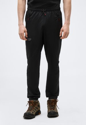 Mężczyzna ubrany w czarne spodnie dresowe z elastycznym pasem i zamykanymi kieszeniami, w połączeniu z brązowo-zielonymi butami trekkingowymi.