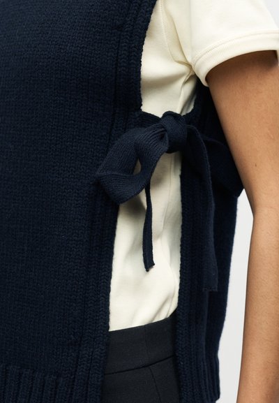 Πλεκτό γιλέκο σε ναυτικό μπλε με υφή, με κλείσιμο στο πλάι με κορδόνι, φορεμένο πάνω από κρεμ πουκάμισο και μαύρο παντελόνι με ψηλή μέση.