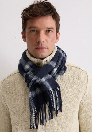 Navy blue plaid scarf made of soft fabric, featuring white and light gray checks, with fringed edges, worn over a cream-colored knit sweater.