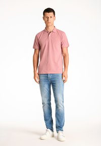 Maglietta polo rosa con maniche corte, colletto piatto e logo sul petto. Indossata con jeans azzurri chiari e sneakers bianche, in piedi contro uno sfondo semplice.