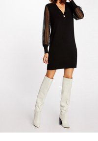 Robe en maille noire à col en V, avec des manches bouffantes transparentes. Un bouton décoratif orne le col. Portée avec des bottes blanches montantes jusqu'au genou.