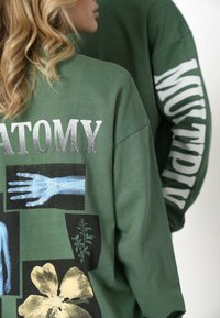 Grøn, oversized sweatshirt med blandede grafiske tryk, herunder anatomiske designs og blomstermotiver. Har store hvide bogstaver på ærmerne.