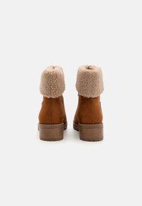 Brune ruskind ankelsko med en fluffy beige kant. Har en chunky hæl og sål i tekstureret brunt for greb. Enkel, afrundet design.