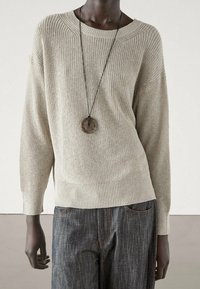 Un pull en maille beige clair avec un décolleté large, des épaules tombantes et des finitions côtelées, associé à un pantalon en denim foncé et un collier avec un pendentif circulaire.