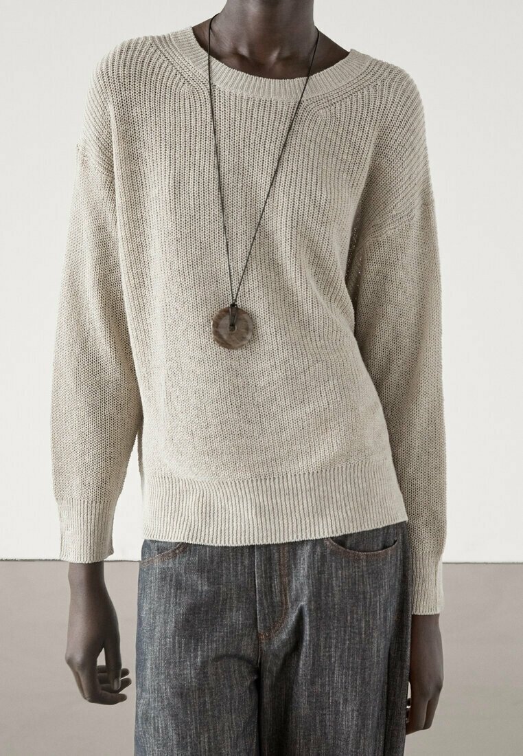 Un pull en maille beige clair avec un décolleté large, des épaules tombantes et des finitions côtelées, associé à un pantalon en denim foncé et un collier avec un pendentif circulaire.
