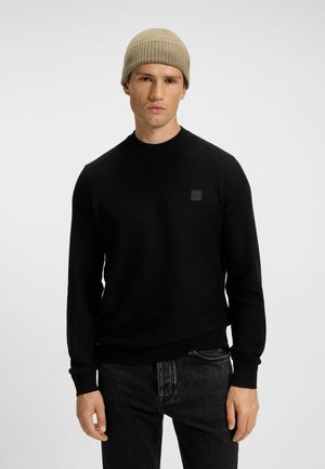 Pull en maille noir avec un col rond, présentant des poignets et un ourlet côtelés, ainsi qu'un petit patch logo noir sur la poitrine. Porté avec un bonnet beige.