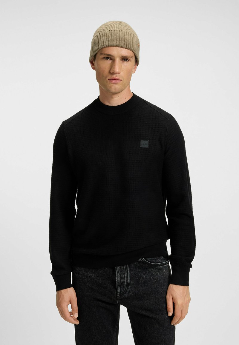 Suéter negro texturizado con cuello redondo, con puños y dobladillo acanalados, y un pequeño parche de logo negro en el pecho. Llevado con un gorro beige.