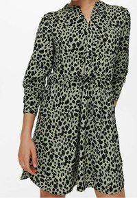 Knapp-up klänning i olivgrön med svart leopardmönster. Har långa ärmar och en midjeskärp, tillverkad av ett lätt tyg.