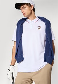 White polo shirt with red and blue trim, featuring a colorful embroidered character. Navy blue sweater draped over shoulders, beige pants, and golf glove.