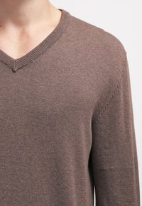 Pull marron à col en V en matière tricotée douce. Il présente un col et des coutures côtelés, avec des manches longues et une silhouette ajustée.