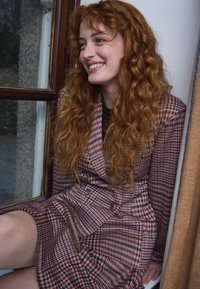 Jeune femme aux longs cheveux roux bouclés sourit assise près de la fenêtre, portant une robe blazer à carreaux avec des boutons.