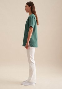 Maglietta verde a maniche corte con tasca, abbinata a pantaloni bianchi a zampa d'elefante e sneaker bianchi. Il tessuto sembra liscio con una vestibilità comoda.