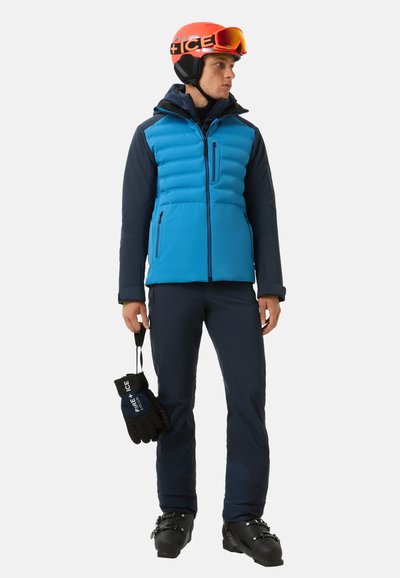 Niebieska kurtka puchowa z granatowymi rękawami, zapięciem na zamek oraz dwoma przednimi kieszeniami. Model ma na sobie rękawice narciarskie i buty, a także pomarańczowy kask narciarski.