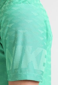 Camiseta deportiva verde claro hecha de un tejido texturizado que presenta un patrón de triángulos repetido y un gran logo impreso en la parte delantera.