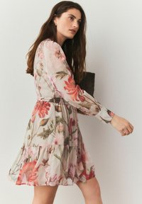 Robe florale en tissu transparent, ornée de fleurs roses et rouges, aux manches longues, avec une taille cintrée et une jupe à volants. Associée à un sac tissé sombre.
