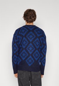 Stickad tröja i mörk marinblått och blått med diamantmönster, ribbad nederkant och en avslappnad passform. Mjuk textur med tydliga geometriska mönster.