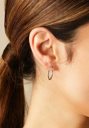 Nahaufnahme eines Ohres einer Frau, die einen kleinen silbernen Hoop-Ohrring trägt, mit braunen Haaren, die zu einem tiefen Pferdeschwanz gebunden sind.