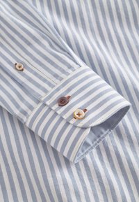 Polsino di camicia a righe blu chiaro e bianche con tre bottoni: due marroni e uno avorio. Tessuto liscio con una leggera lucentezza.