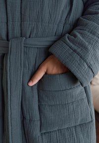 Gesteppter dunkelgrauer Bademantel mit Gurt, aus strukturiertem Stoff und mit seitlicher Tasche, hervorgehoben durch weiches, zerknittertes Material.