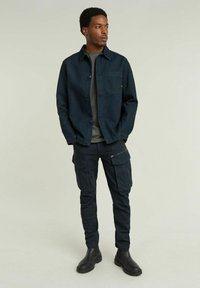 Ciemnoniebieska koszula z guzikami, szara koszulka pod spód, spodnie cargo z kieszeniami na suwak oraz czarne buty za kostkę. Gładka tkanina i dopasowany krój.