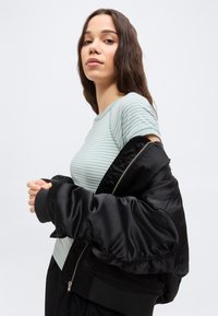 Νέα γυναίκα με μακριά καστανά μαλλιά που φοράει ένα πράσινο ριγέ πουκάμισο και μια μαύρη ζακέτα ριγμένη στους ώμους της, μπροστά σε ένα απλό φόντο.