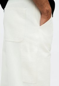 Χέρι που ακουμπά στην πλαϊνή τσέπη ενός λευκού παντελονιού με μεγάλες ορθογώνιες επιδιορθωτικές τσέπες σε απλό φόντο.