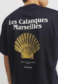 Černé bavlněné tričko s bílým nápisem "Les Calanques Marseilles" nad žlutým motivem mušle na zadní straně, volný střih.
