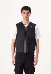 Svart ermeløs vest med to frontlommer, gul glidelås og logoaksent. Båret over en hvit t-skjorte, med jevn tekstur og lett design.