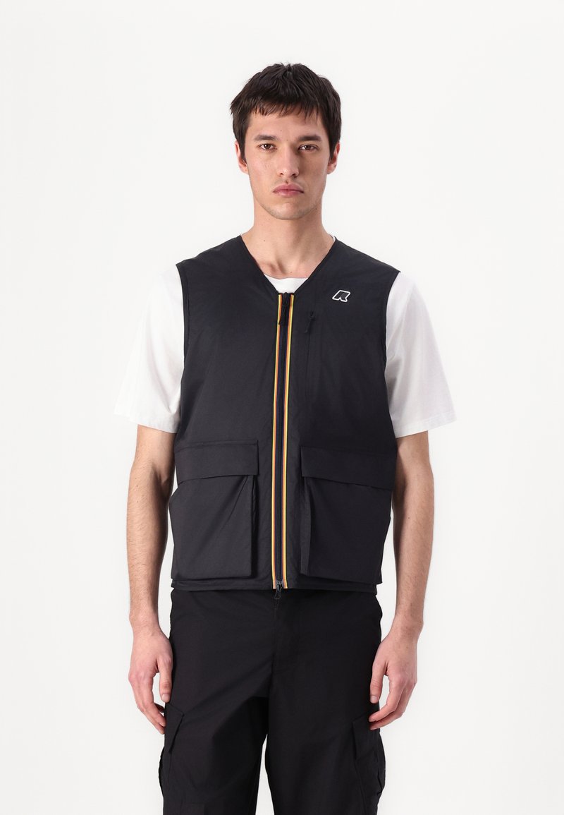 Svart ermeløs vest med to frontlommer, gul glidelås og logoaksent. Båret over en hvit t-skjorte, med jevn tekstur og lett design.