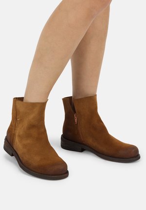 Braune Wildleder-Stiefeletten mit seitlichen Reißverschlüssen, getragen an nackten Beinen vor einfachem Hintergrund.