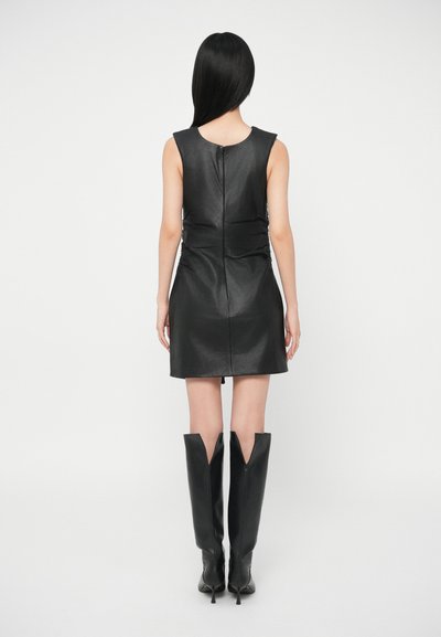 Fekete bőrruha testhezálló szabással, magas nyakkivágással, hátul látható cipzárral, és magas, hegyes orrú fekete csizmákkal párosítva.