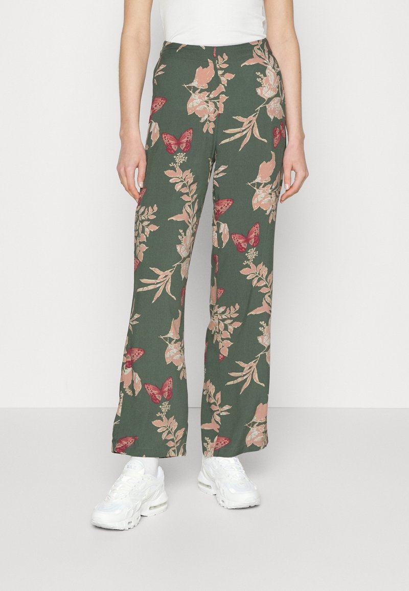 Pantalon vert à jambes larges avec motif floral rose et papillon rouge, porté avec des baskets blanches et un haut blanc.