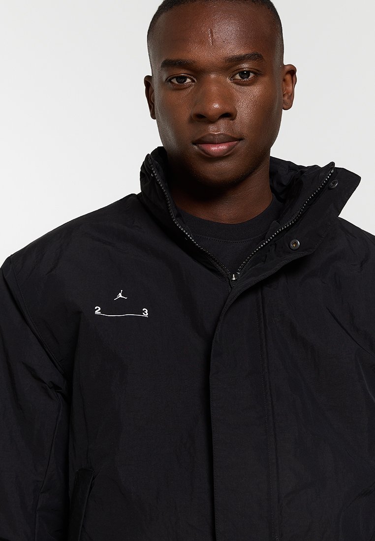 Joven con una chaqueta negra con cremallera y un logo minimalista blanco que presenta una pequeña figura de Jumpman y los números 2 y 3 en el pecho.
