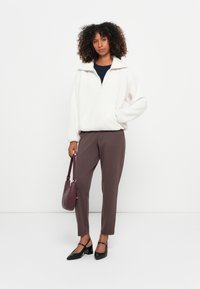 Witte fleece trui met rits tot de helft en staande kraag, gecombineerd met bruine tapered broeken en zwarte puntige hakken, met een bordeauxkleurige tas.