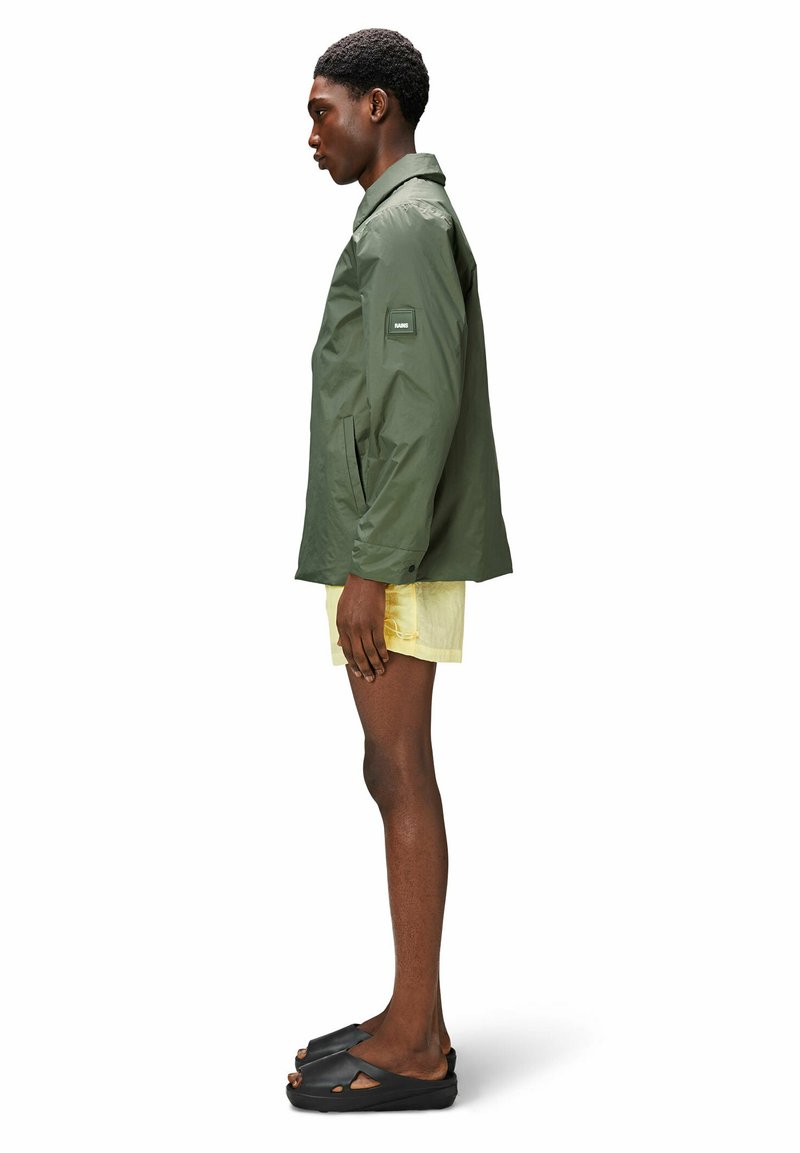 Veste imperméable olive avec un col, des poches latérales et un patch de marque sur la manche, portée avec un short jaune et des chaussures noires.