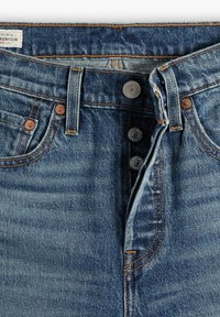 Jean en denim bleu devant avec braguette à boutons visible, passants de ceinture, rivets en cuivre et vue partielle d'une étiquette de marque blanche sur la ceinture.