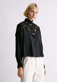 Blusa nera con colletto arricciato, con dettagli in pizzo sullo scollo e maniche lunghe con polsini arricciati. Indossata con pantaloni bianchi.