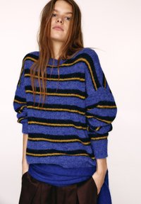 Modrý pruhovaný sveter so žltými a čiernymi vodorovnými pruhmi, vyrobený z textúrovaného úpletu. Dizajn má pohodlný strih a spustené ramená.