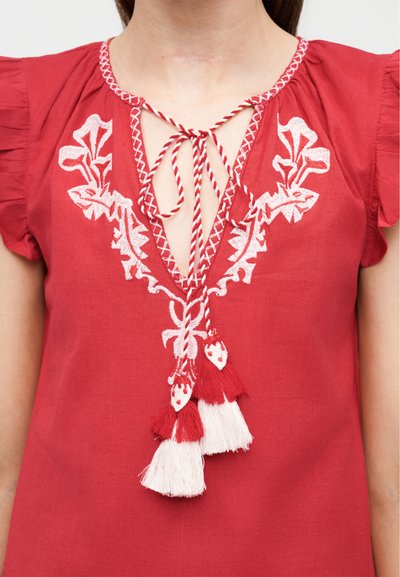 Κόκκινη μπλούζα με λευκή κεντητή διακόσμηση λουλουδιών γύρω από το ντεκολτέ, δεμένη με στριμμένο κόκκινο-λευκό κορδόνι που καταλήγει σε κόκκινες και λευκές φούντες.