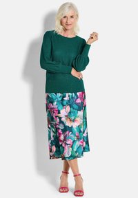 Dunkelgrüner Pullover aus strukturiertem Garn, kombiniert mit einem Midi-Rock mit Blumenmuster, der pinke und türkisfarbene Blumen zeigt, sowie pinken Sandalen mit Riemchen.