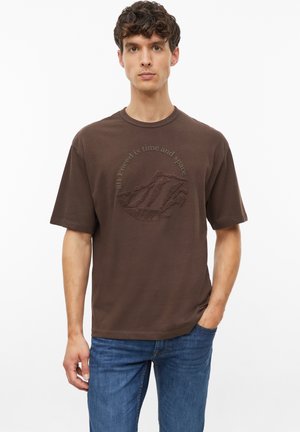 Junger Mann mit lockigem Haar trägt ein braunes T-Shirt mit texturiertem Bergmotiv und Schriftzug, kombiniert mit blauen Jeans, steht vor weißem Hintergrund.