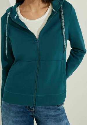 Femme portant un sweat à capuche zippé bleu sarcelle avec des cordons imprimés et un t-shirt blanc en dessous, vêtue d'un jean bleu, debout avec une main derrière le dos.