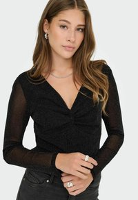 Giovane donna con capelli mossi sciolti, indossa un top nero scintillante a maniche lunghe, una collana a catena d'argento, orecchini a cerchio e anelli d'argento su entrambe le mani.