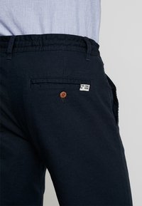 Pantaloni di cotone blu scuro con vita elasticizzata, dotati di una tasca posteriore con un bottone marrone e un'etichetta bianca con la scritta "THE CODE WEAR."